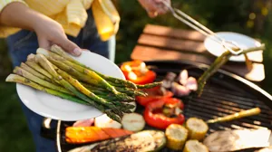 En hand håller i en tallrik med grillad sparris, i bakgrunden skymtas grönsaker på en klotgrill.