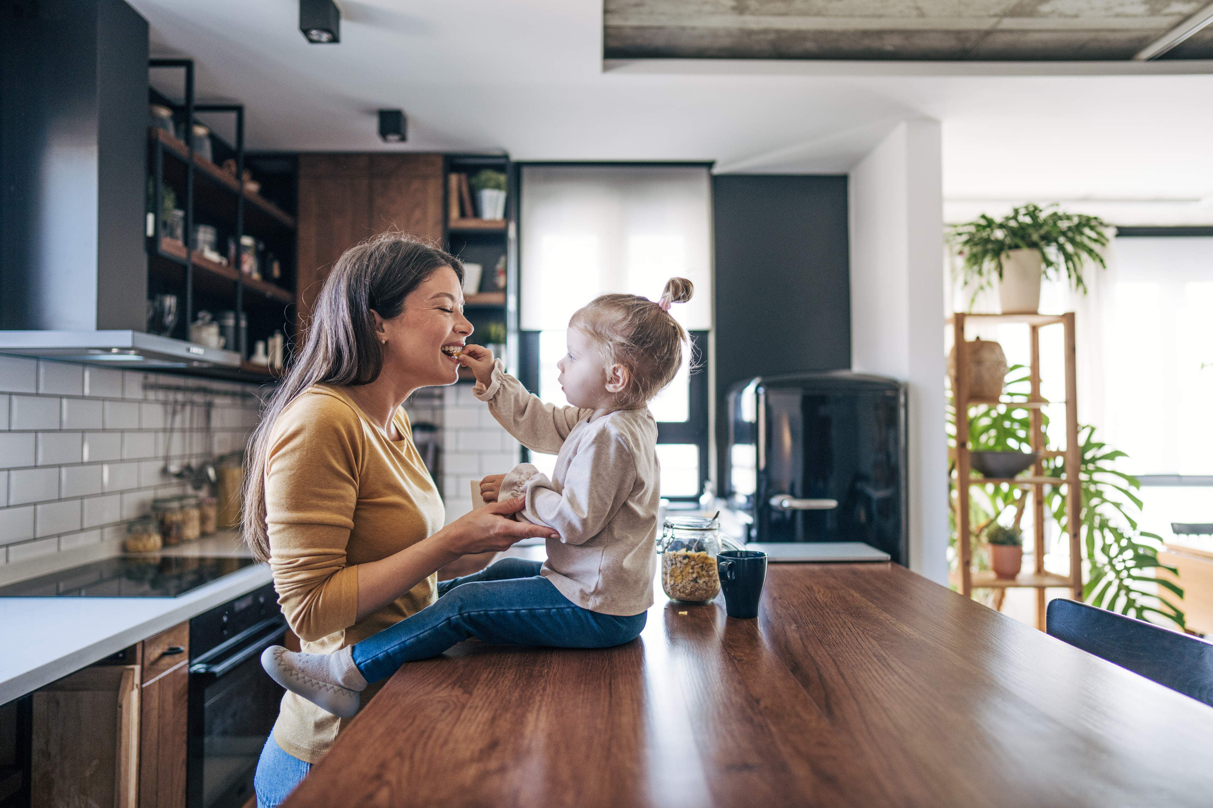 Ett barn sitter på en köksbänk i ett modernt kök och matar en leende kvinna som står framför bänken.