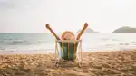 Bild på person som sitter i en solstol på en strand framför ett hav med händer utsträckta