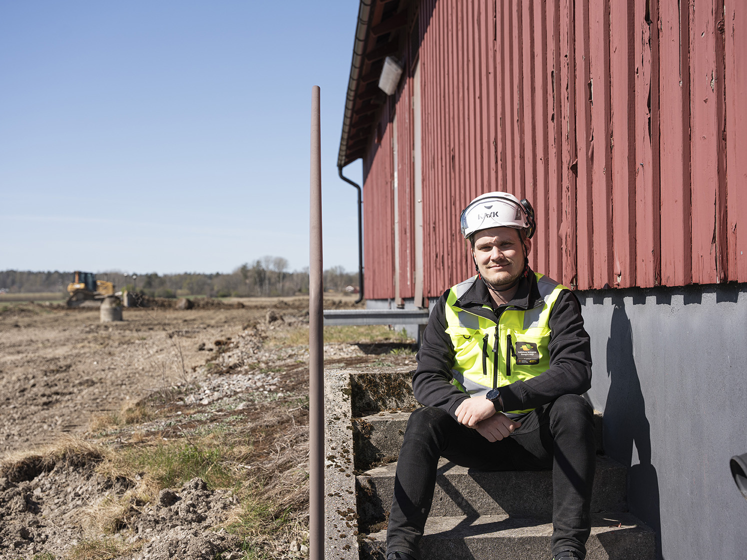 Projektledaren Jimmy sitter på en trappa iklädd varselväst och hjälm. I bakgrunden på ett fält syns en grävmaskin.