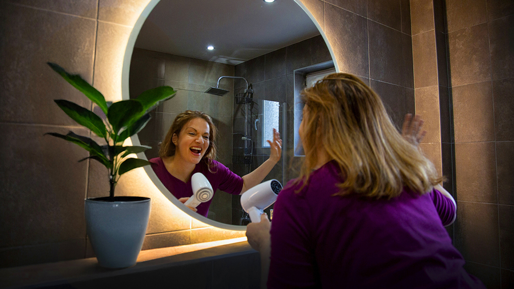 Sjungande kvinna framför badrumsspegel använder hårfön som mikrofon.