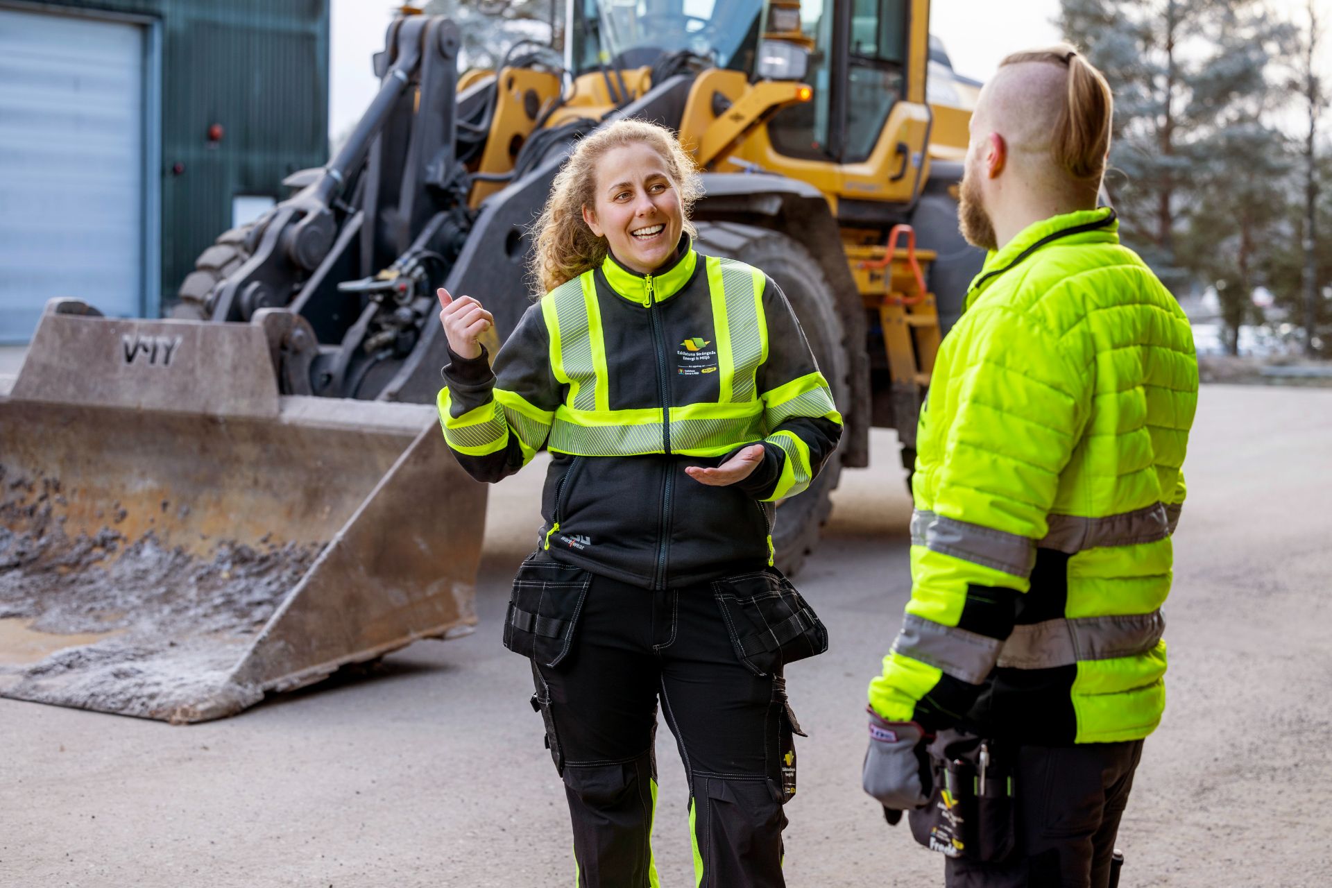 Två personer står framför en grävmaskin och pratar med varandra.