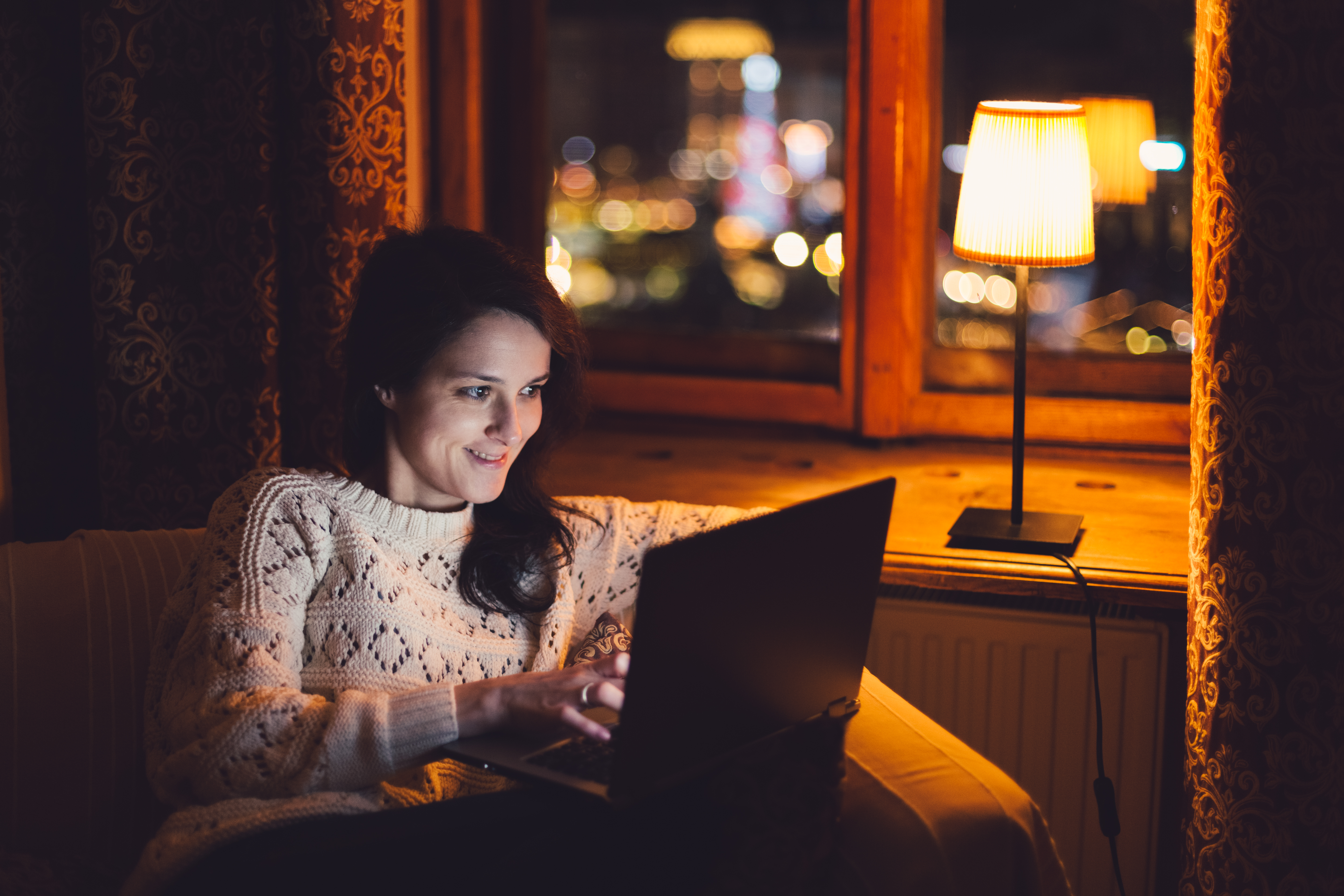 Kvinna som sitter i en fåtölj vid ett upplyst fönster med en dator i knät