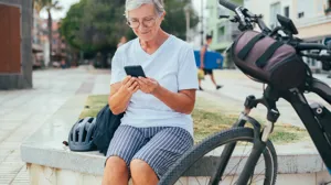 En äldre kvinna som sitter på en bänk bredvid en cykel och tittar i sin telefon.